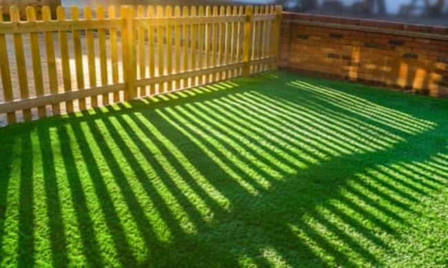 Shadows of a wooden picket fence in a front yard, front garden with artificial grass as a lawn and a red brick perimeter wall.